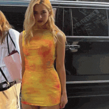 a woman in a yellow tie dye dress stands in front of a black van that says dsquared on the side