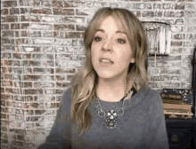a woman is standing in front of a brick wall wearing a necklace and a sweater .