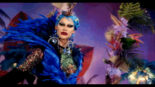 a drag queen with blue hair and feathers stands in front of flowers