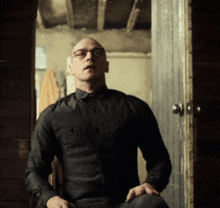 a bald man wearing glasses and a black shirt is sitting in front of a door