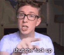 a man wearing glasses is pointing at the camera and saying shut the fuck up .