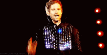 a man is singing into a microphone on a stage while wearing a light up vest .