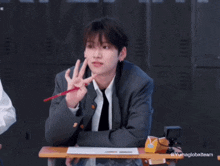 a young man in a suit and tie is sitting at a desk and making a peace sign