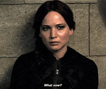 a woman in a black jacket says " what now " in front of a brick wall