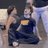 a man is sitting on the floor with his mouth open and another man is standing behind him .
