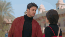 a man in a red jacket and a woman in a black turtleneck are looking at each other .