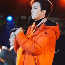 a man in an orange jacket is standing on a stage with a microphone in his hand .