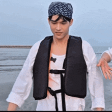 a man wearing a life jacket and a bandana stands on the beach