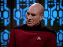 a bald man wearing a red sweater with a silver star trek badge