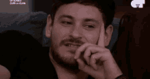 a man with a beard is smiling while sitting on a couch with his hand on his chin .