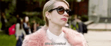 a woman wearing sunglasses and a pink fur coat is standing in front of a crowd .