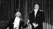 a black and white photo of two men in tuxedos and top hats standing on a stage .