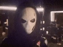 a person wearing a white mask and a black hoodie is standing in a dark room .