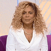 a woman with curly blonde hair is sitting on a purple couch wearing a white jacket and gold necklace .