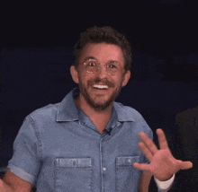 a man wearing glasses and a denim shirt smiles and waves his hand