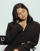 a woman in a black jacket sits in front of a sign that says ' lc '