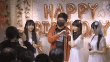 a man wearing a mask stands in front of balloons that spell out happy birthday