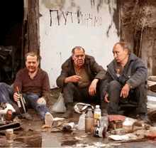 three men are sitting on the ground in front of a wall that says " putin "