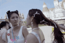 two women are standing next to each other in front of a castle . one has a bow in her hair .