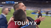 a man is hugging another man on a soccer field with the words tounyx on the screen .