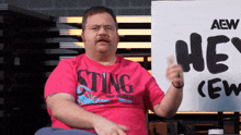 a man wearing a pink shirt that says sting is sitting in front of a sign that says he 's crew