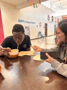 a boy and a girl are sitting at a table eating food in front of a wall that says 100 % college bound