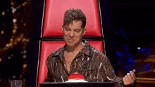 a man with curly hair is sitting in a red chair with a red button in front of him .