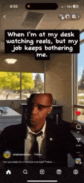 a man in a suit and tie is sitting at a desk watching reels on a cell phone .