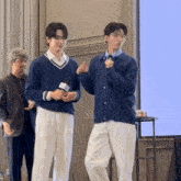 two men are standing next to each other and one is holding a microphone and the other is giving a thumbs up