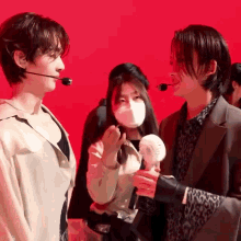 two men are standing next to each other in front of a red background while a woman wearing a mask holds a fan .