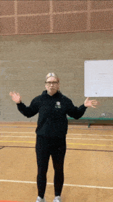 a woman wearing glasses and a black hoodie with a shield on it stands in a gym with her arms outstretched