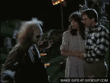 a man and a woman are standing next to each other in a cemetery while a man talks to a zombie .