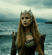 a woman with long red hair and a crown on her head stands in front of the ocean