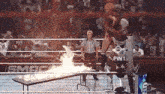 a wrestler is jumping over a table with flames coming out of it while a referee watches .