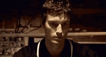 a young man wearing a black shirt and an apron is looking at the camera in a dark room .