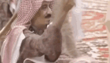 a man in a keffiyeh is standing in front of a wall with his hand up .