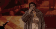 a woman in a fur coat is standing in front of a microphone and raising her hand .