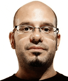 a bald man wearing glasses and a beard looks at the camera