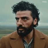 a man with curly hair and a beard is wearing a brown coat and looking at the camera .