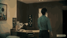 a man in a green shirt is standing in front of a desk with a lamp on it and a sign that says kapwing on it