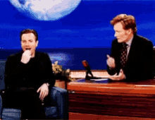 two men are sitting at a table talking to each other with a planet in the background