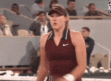 a woman in a maroon nike tank top is standing on a tennis court in front of a crowd of spectators .