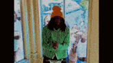 a man wearing a green and white striped shirt and an orange beanie stands in front of a window