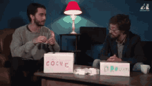 two men sitting at a table with boxes that say coche and drogue