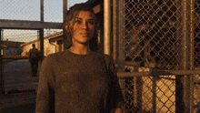 a woman stands in front of a chain link fence with a man walking in the background