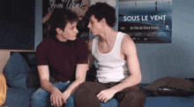 two men are sitting on a bed in front of a sign that says sous le vent