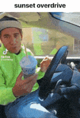 a man in a green hoodie is driving a car and holding a cup of milkshake with a straw ..