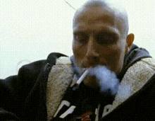 a man with a shaved head is smoking a cigarette while wearing a jacket with the letter d on it .