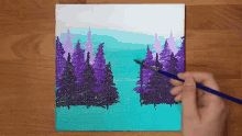 a person is painting a painting of a lake with purple trees on a canvas .