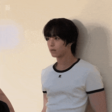 a young man leaning against a wall wearing a white t-shirt with a black letter z on the front